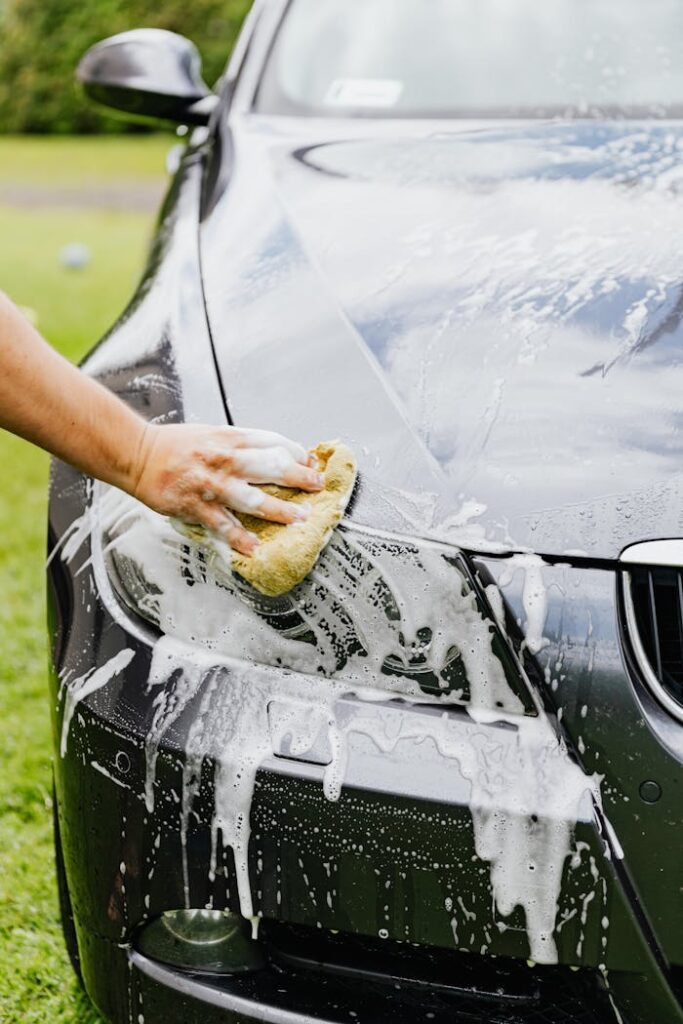 Person washing a black car's headlight with a sponge and soap outdoors.